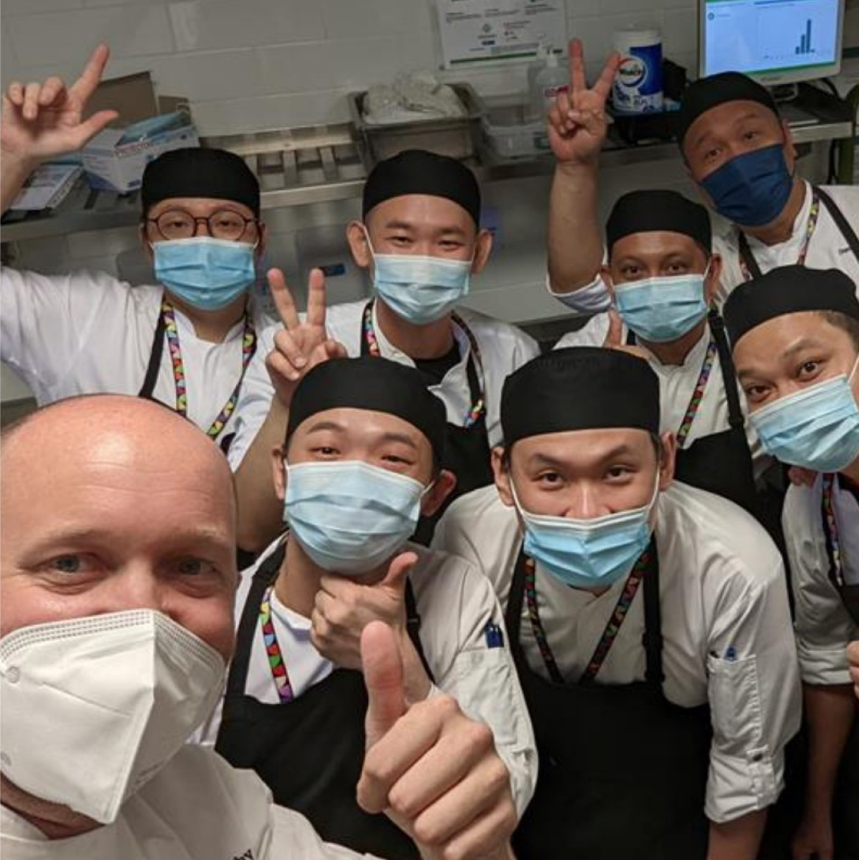 A group of chefs in masks smiling and giving thumbs up, showcasing teamwork and positivity in the kitchen