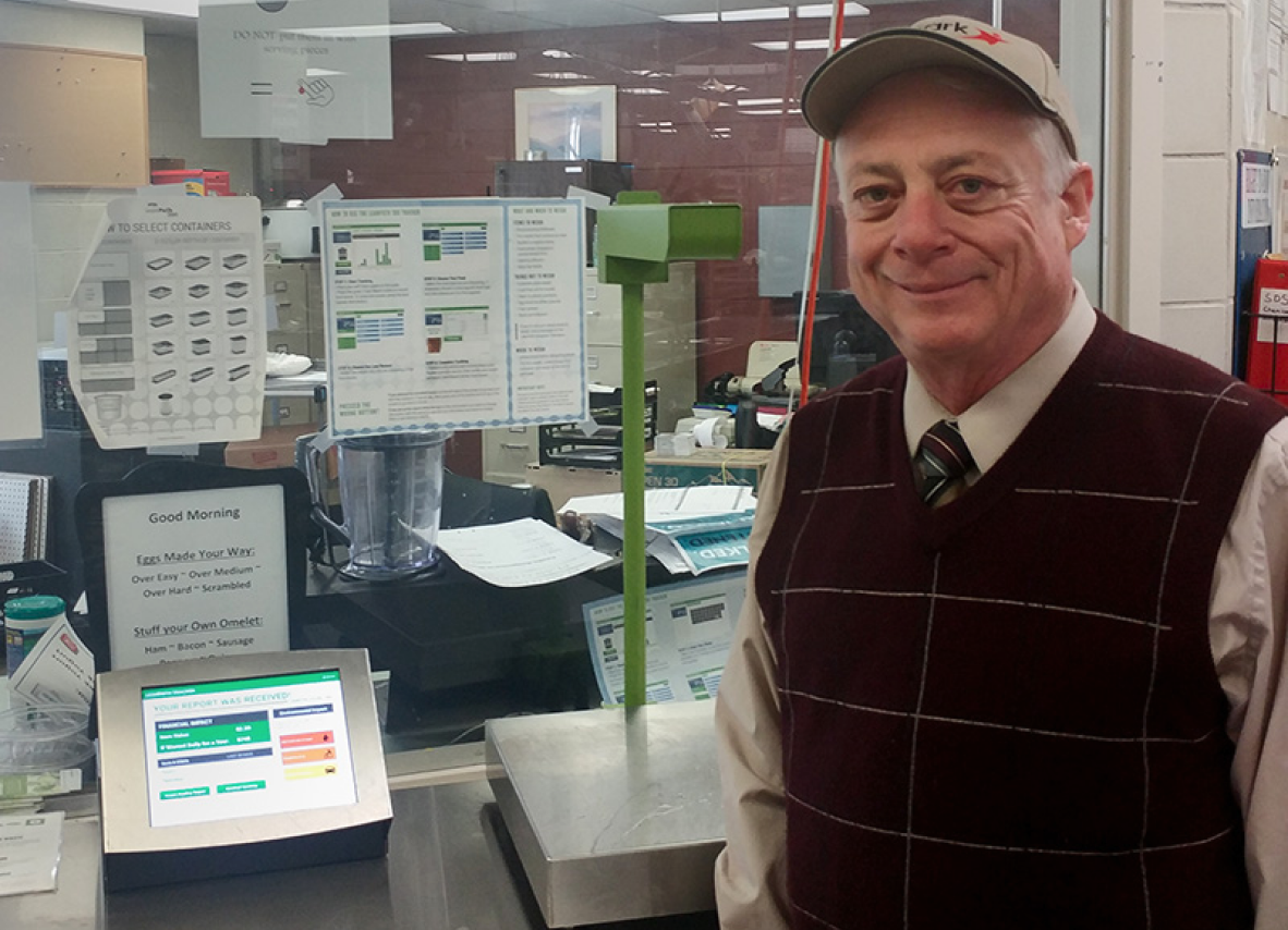 A man wearing a vest and hat stands confidently in front of a computer, engaged in his work environment