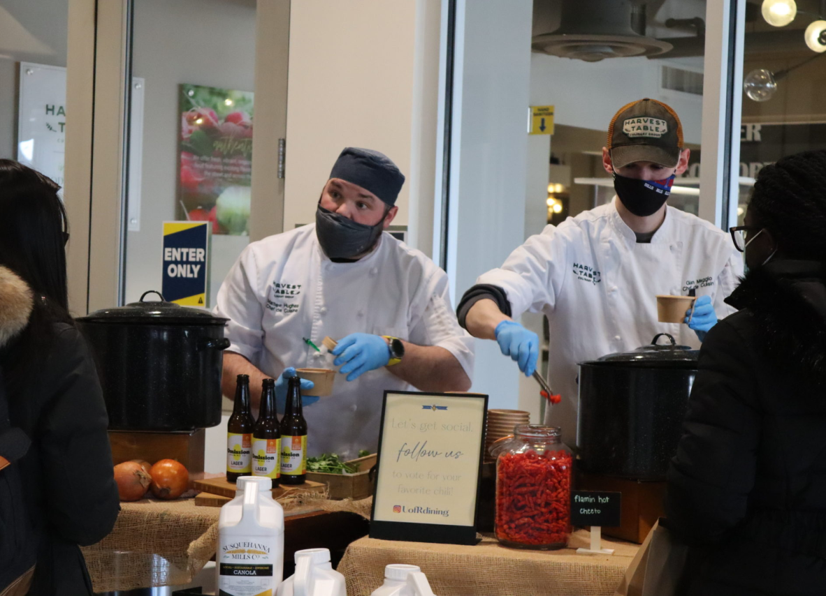 Two individuals wearing masks and gloves are engaged in food preparation in a clean kitchen environment