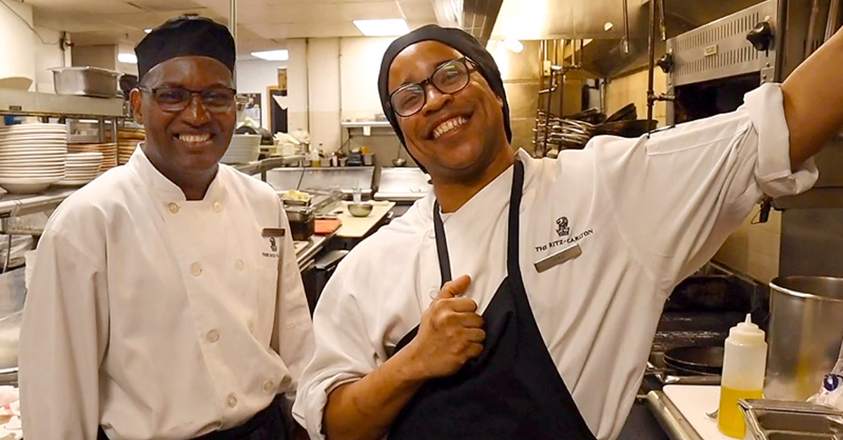 Two chefs smiling in a kitchen, surrounded by pots and pans, showcasing a joyful culinary atmosphere